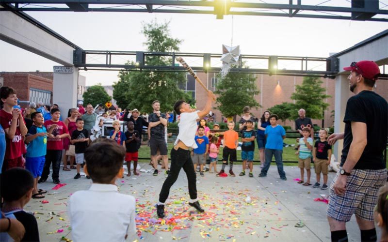 Group of people outdoors, child hitting piñata, colorful confetti on ground, festive atmosphere.