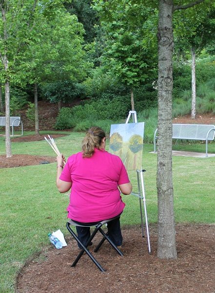 Person in pink shirt painting on easel in park with trees and benches in background.