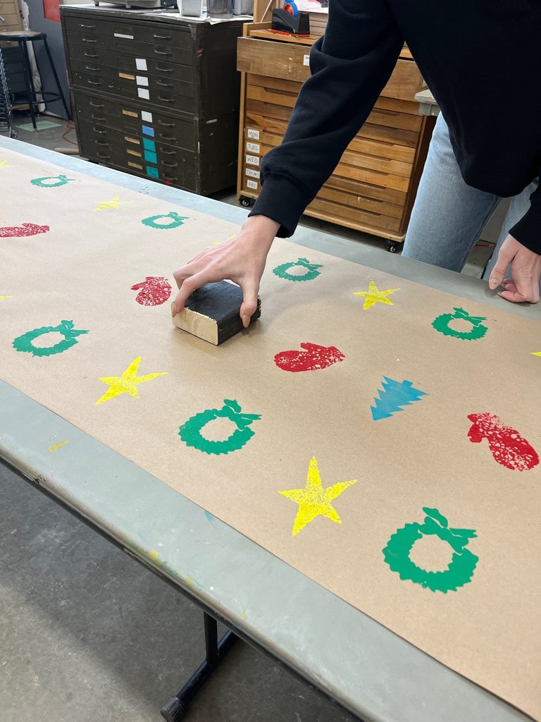 Person stamping holiday-themed patterns on brown paper in an art studio.