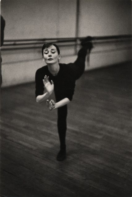 Dancer balances on one leg in dim studio, with extended leg and barre visible.