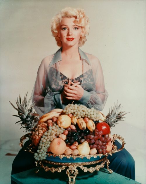 Woman in light blouse with fruit bowl including grapes, pineapples, apples; indoor neutral setting.