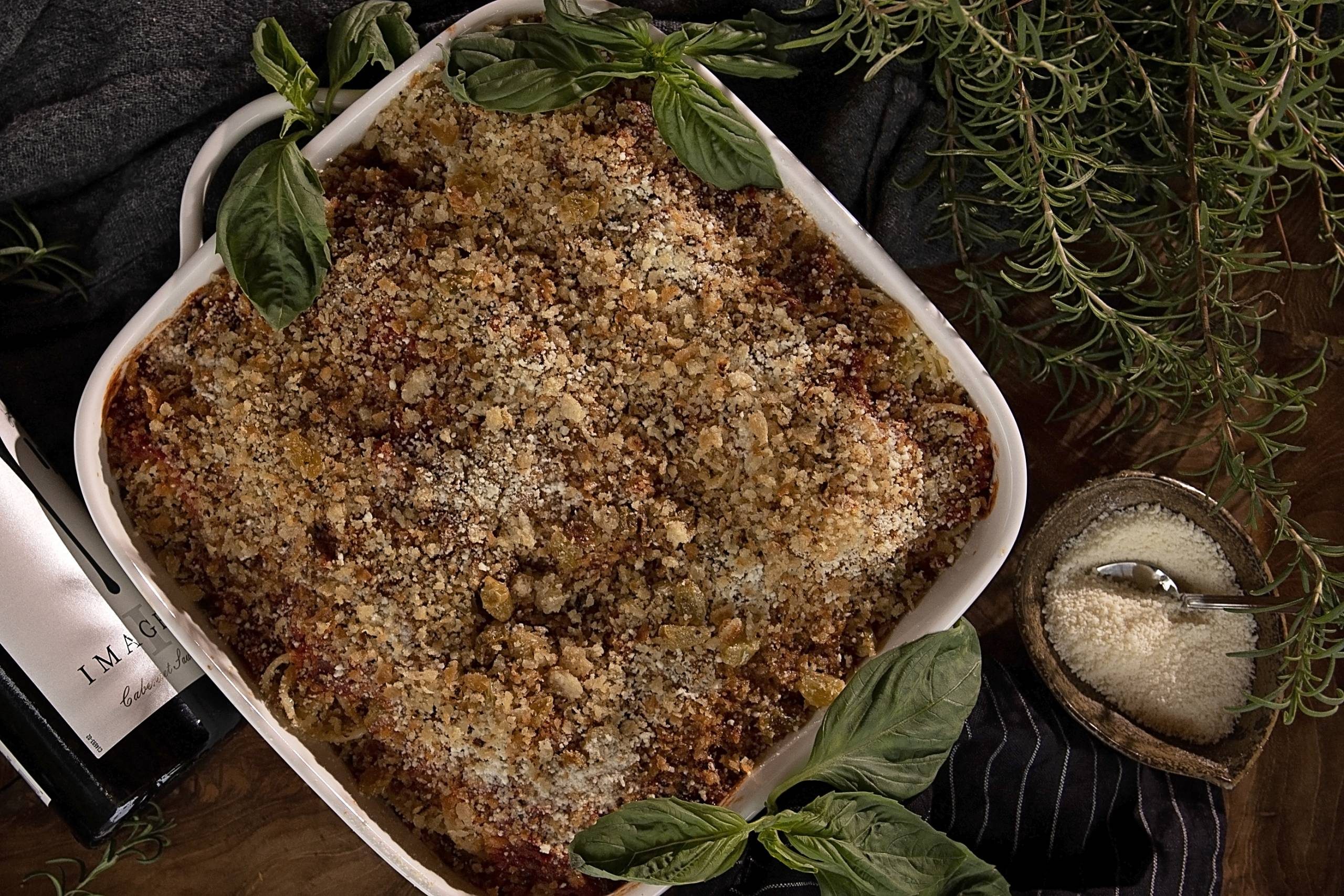 Casserole with breadcrumb, cheese topping, basil; wine bottle, grated cheese, rosemary nearby.
