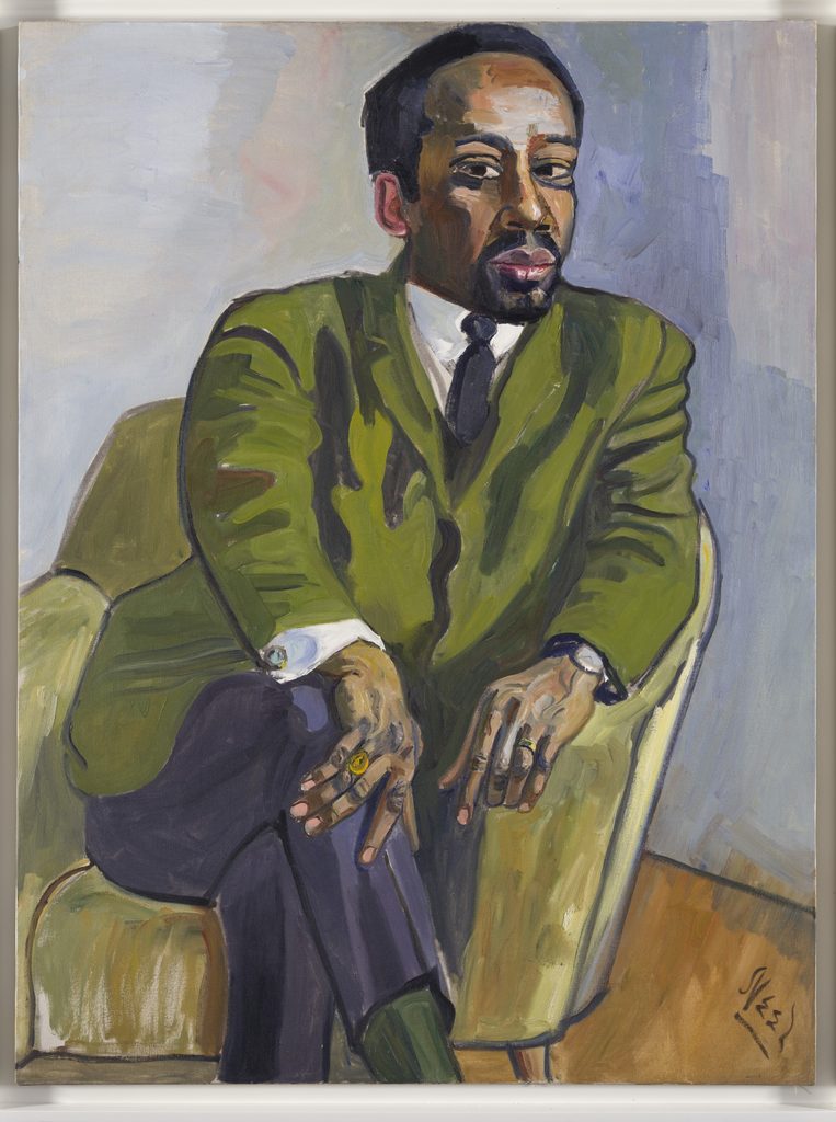 Seated man in green suit with black tie, crossed legs, rings, and muted background.