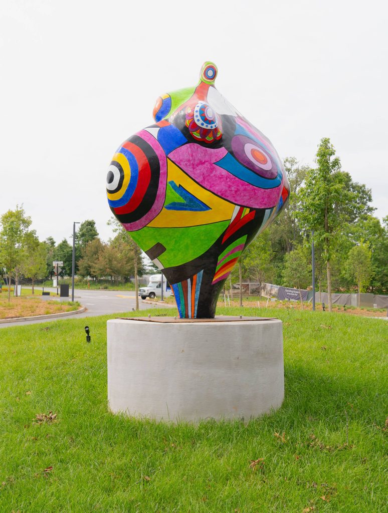 Colorful abstract sculpture with geometric patterns on concrete pedestal in grassy area.