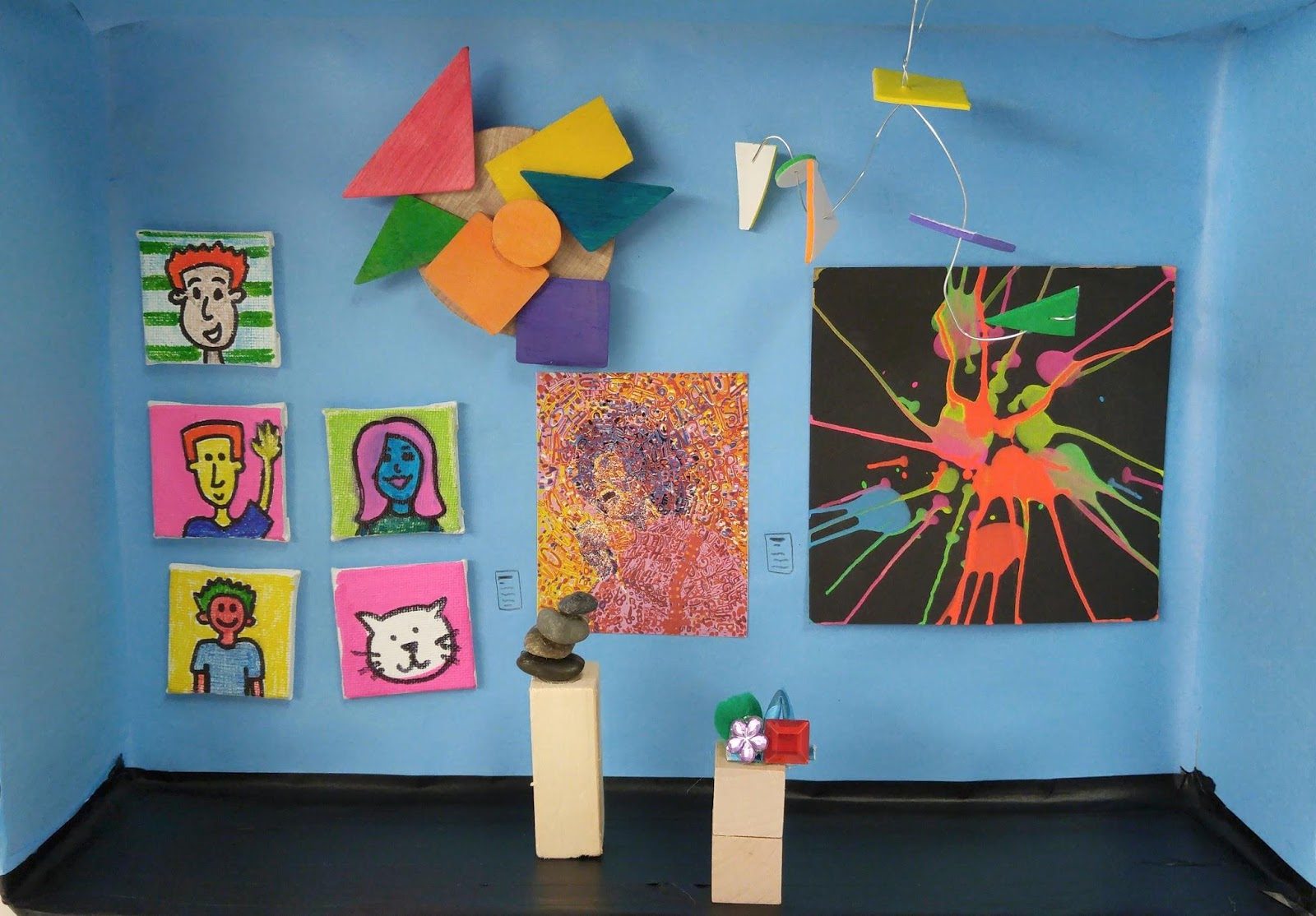Art display with colorful geometric shapes, pop-art portraits, and splatter painting on a blue wall.