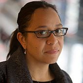 Person with glasses in dark coat looks at camera; blurred background.