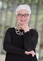 Individual with white hair, glasses, smiling in black outfit and necklace; hand near chin.