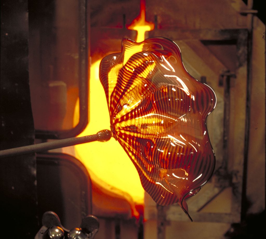 Glassblowing process with a striped glass piece on a rod near a glowing furnace.