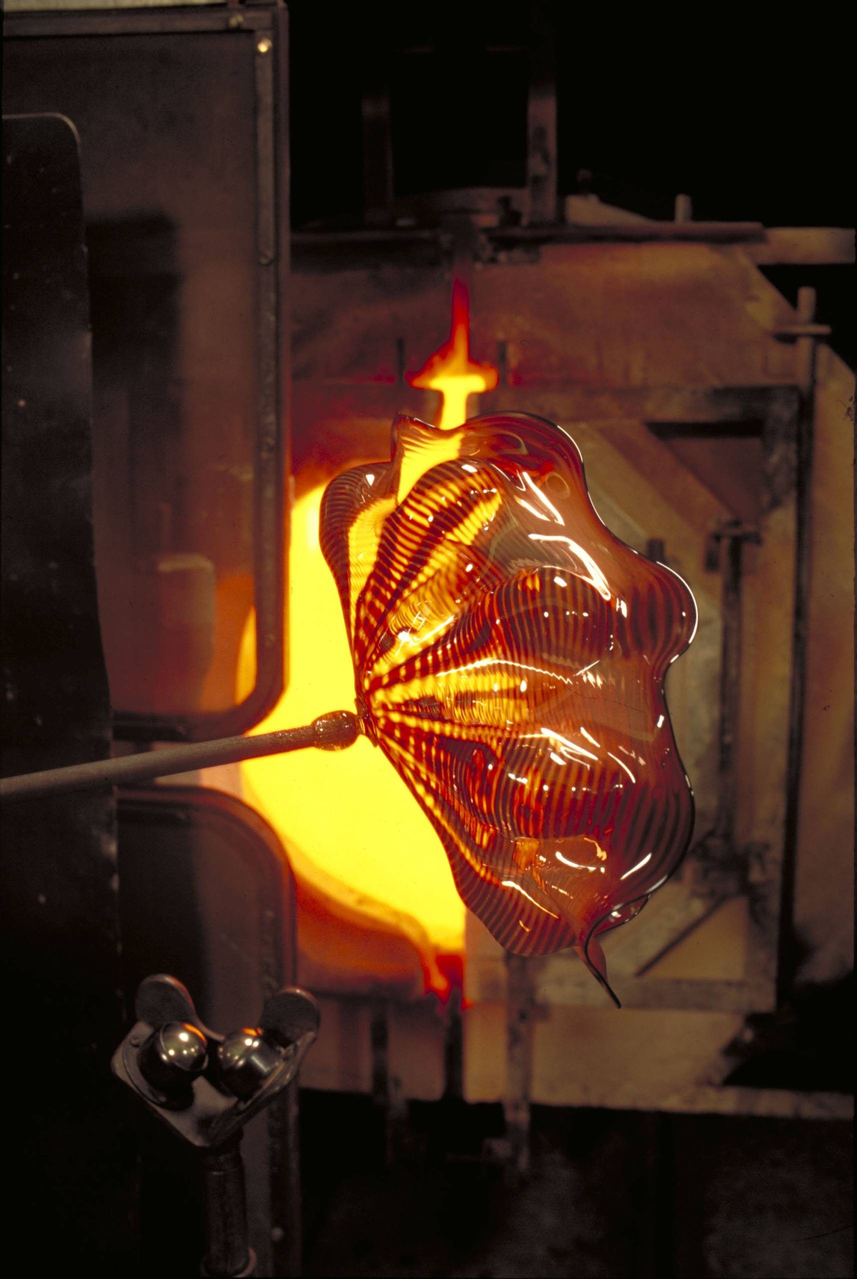 Glowing art glass with wavy pattern being shaped on a rod near a furnace.