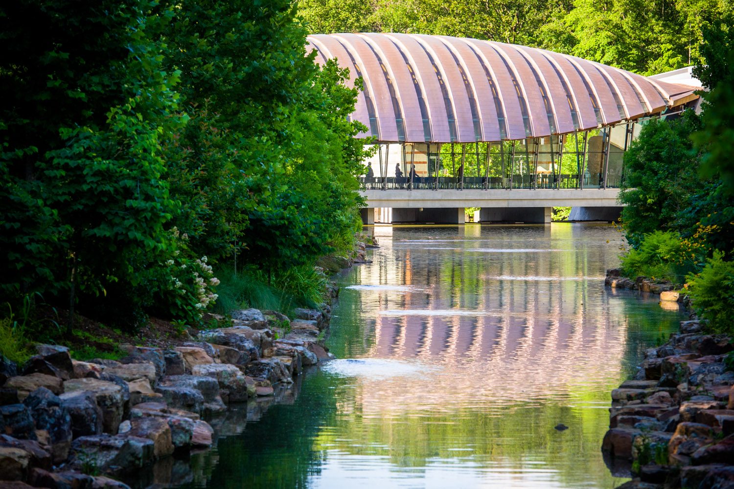 Modern curved-roof building over water, with lush greenery and stone-lined banks.