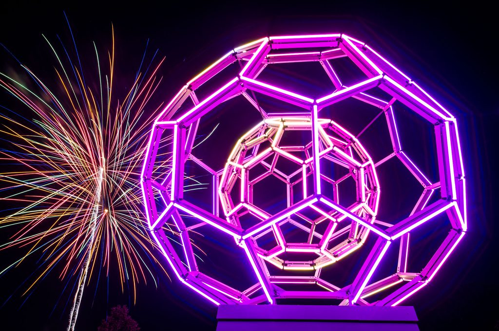 Neon geometric sculpture with pink and purple glow, fireworks burst in the night sky beside it.