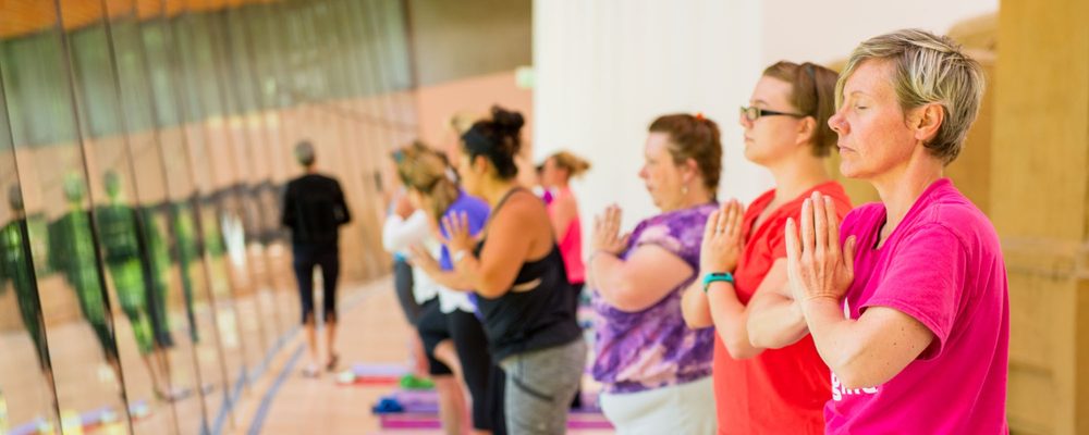 Indoor yoga class with participants standing in prayer position and eyes closed.