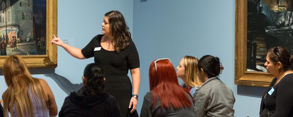 Group in gallery, woman explains painting; street scene and dark painting visible on walls.