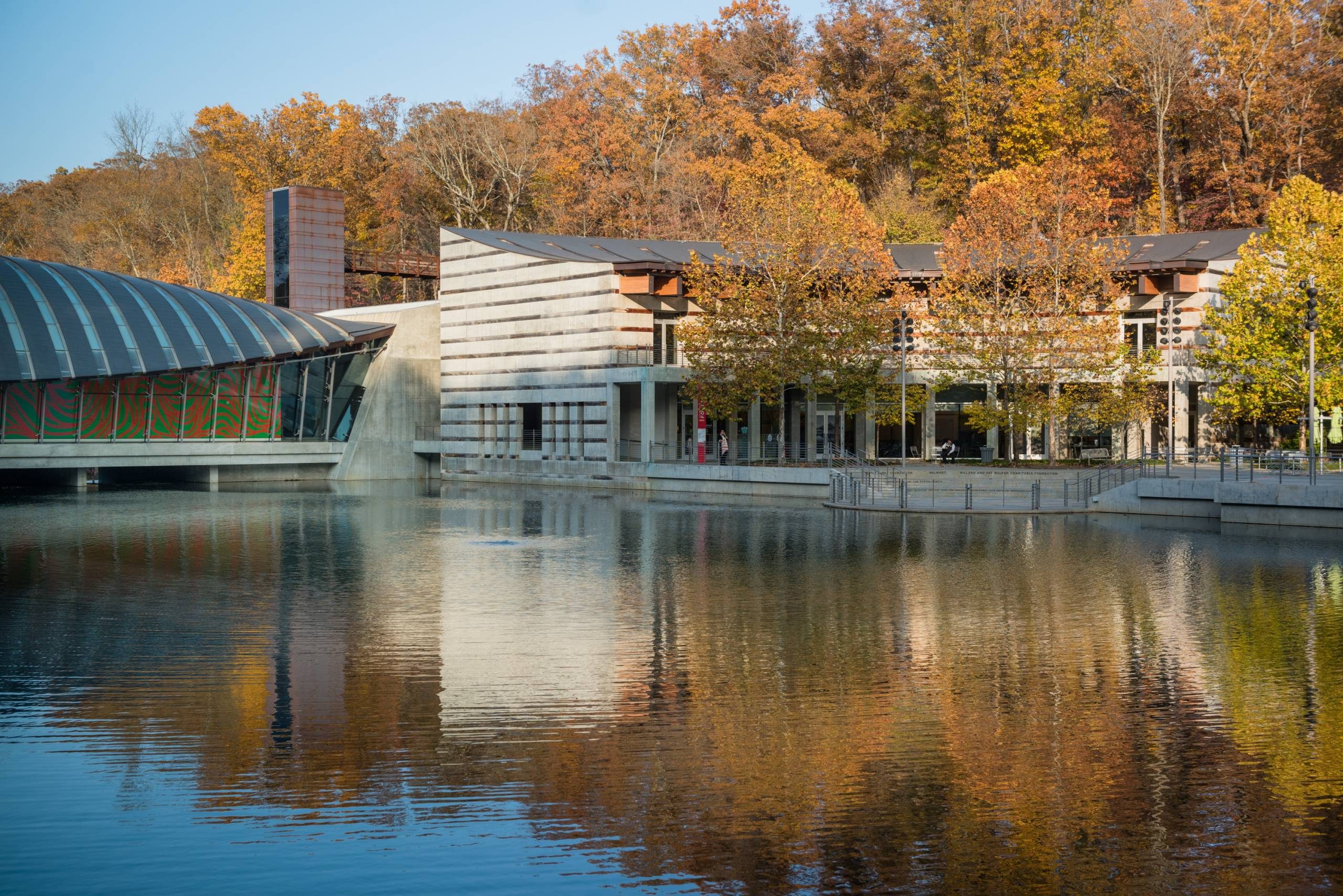 Modern building with large glass windows by a reflective lake, surrounded by autumn trees.