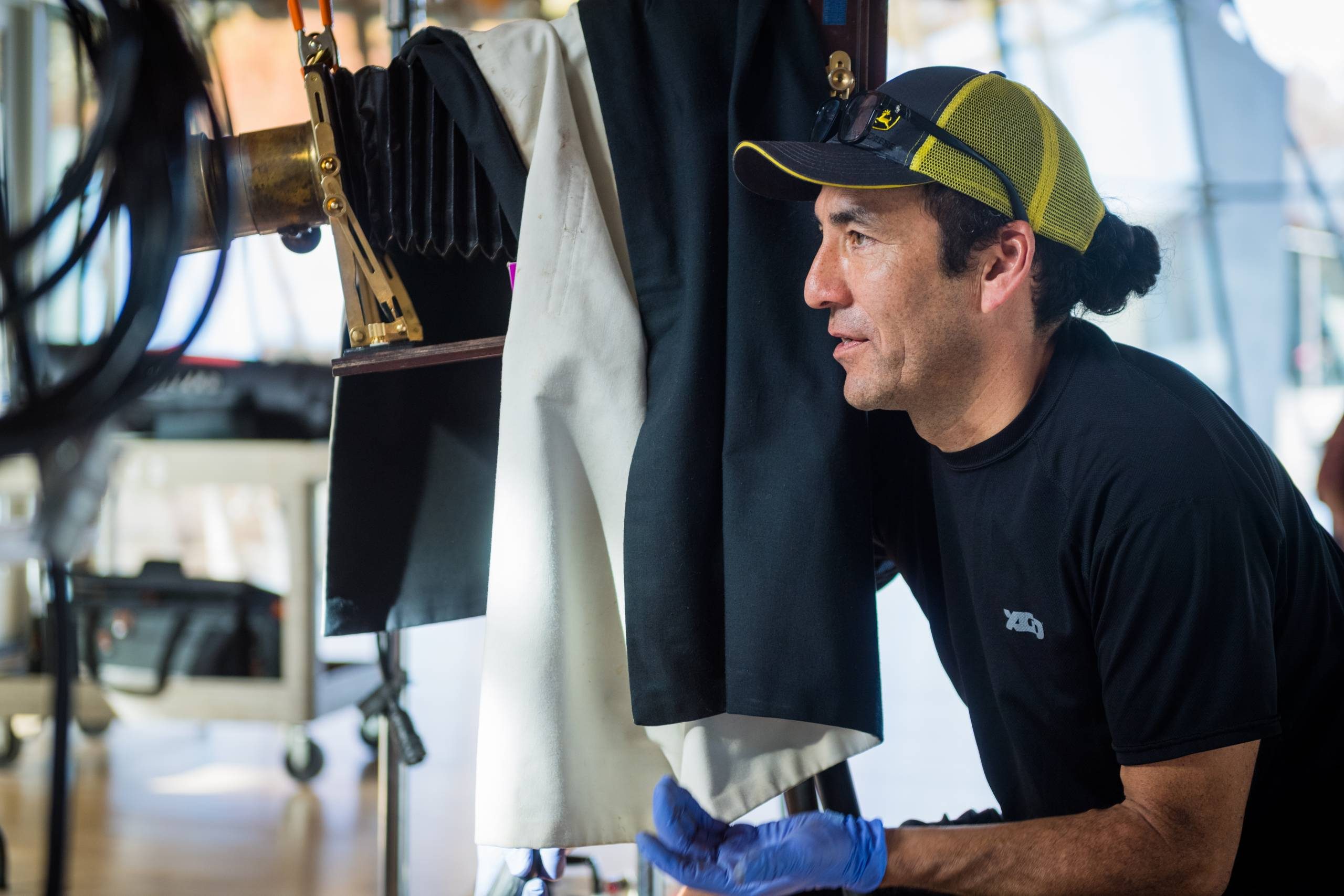 Man in cap and blue gloves working with large camera in a workshop or studio setting.