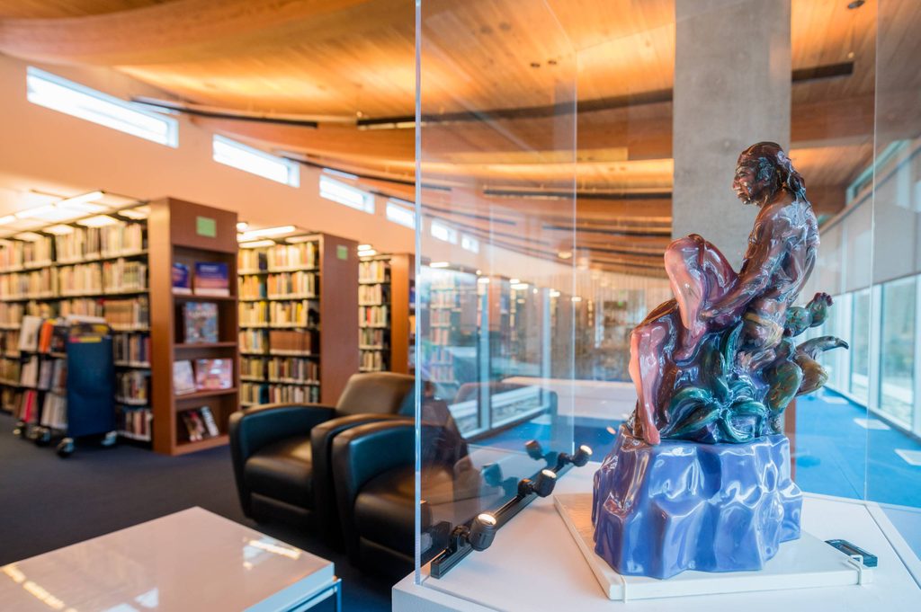 Ceramic sculpture of seated figure in glass case within modern library with bookshelves and armchairs.