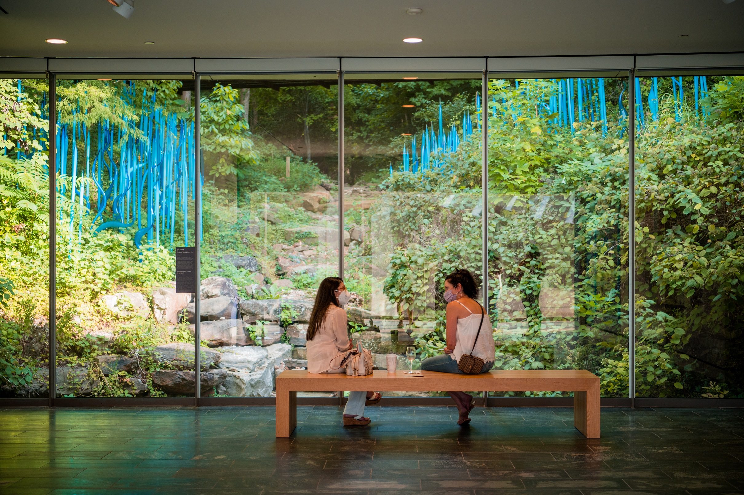 two guests sit on a bench in front of a glass window looking out to a Chihuly artwork