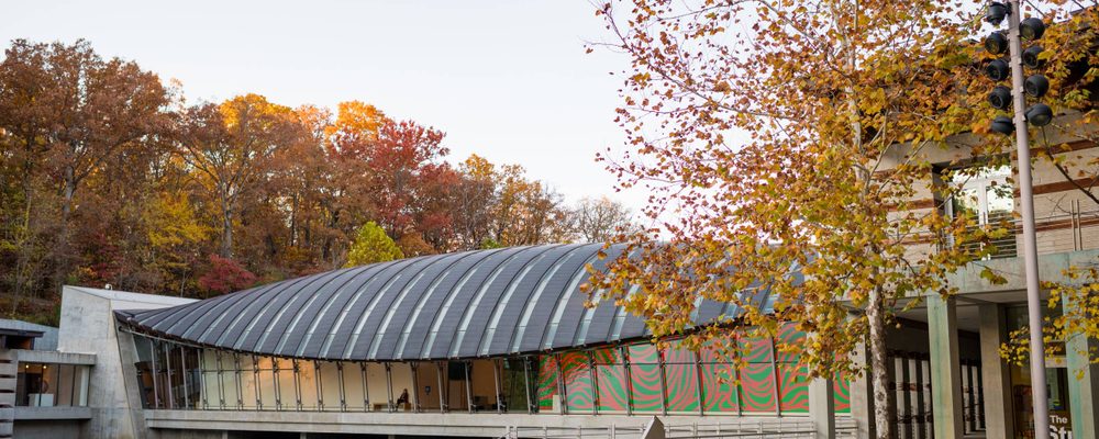 Modern building with curved glass roof next to water and autumn trees with stairs and railings.