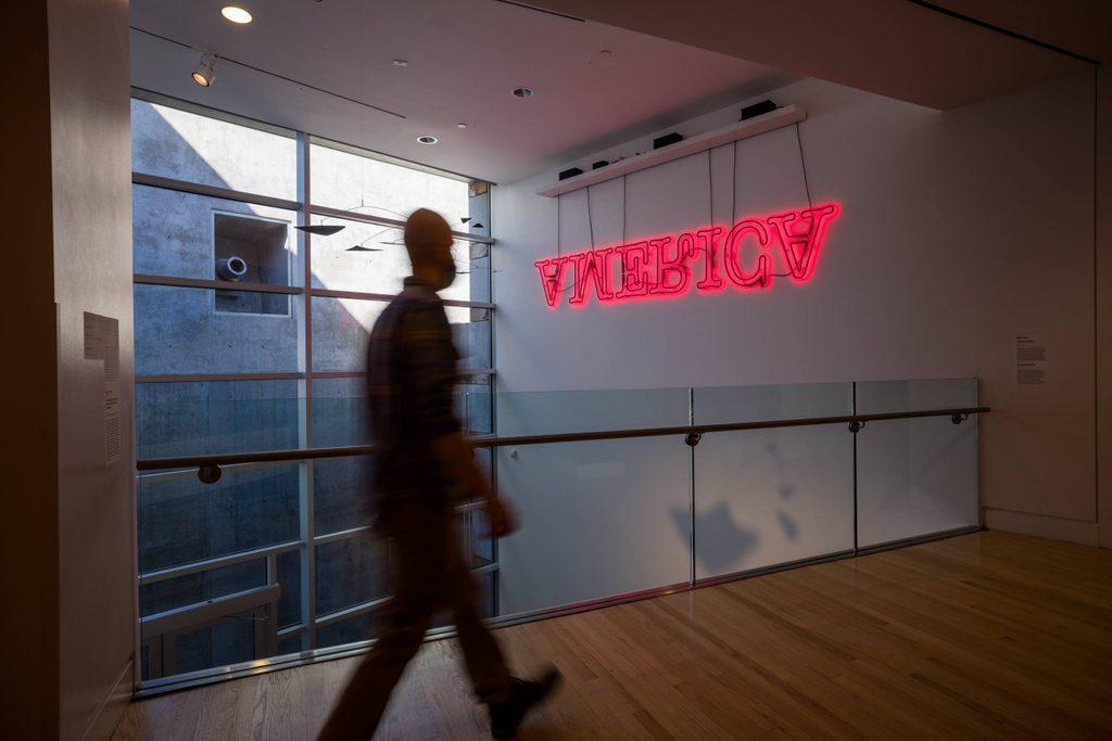 a person walks a gallery bridge looking at a neon-red work on a wall that spells 