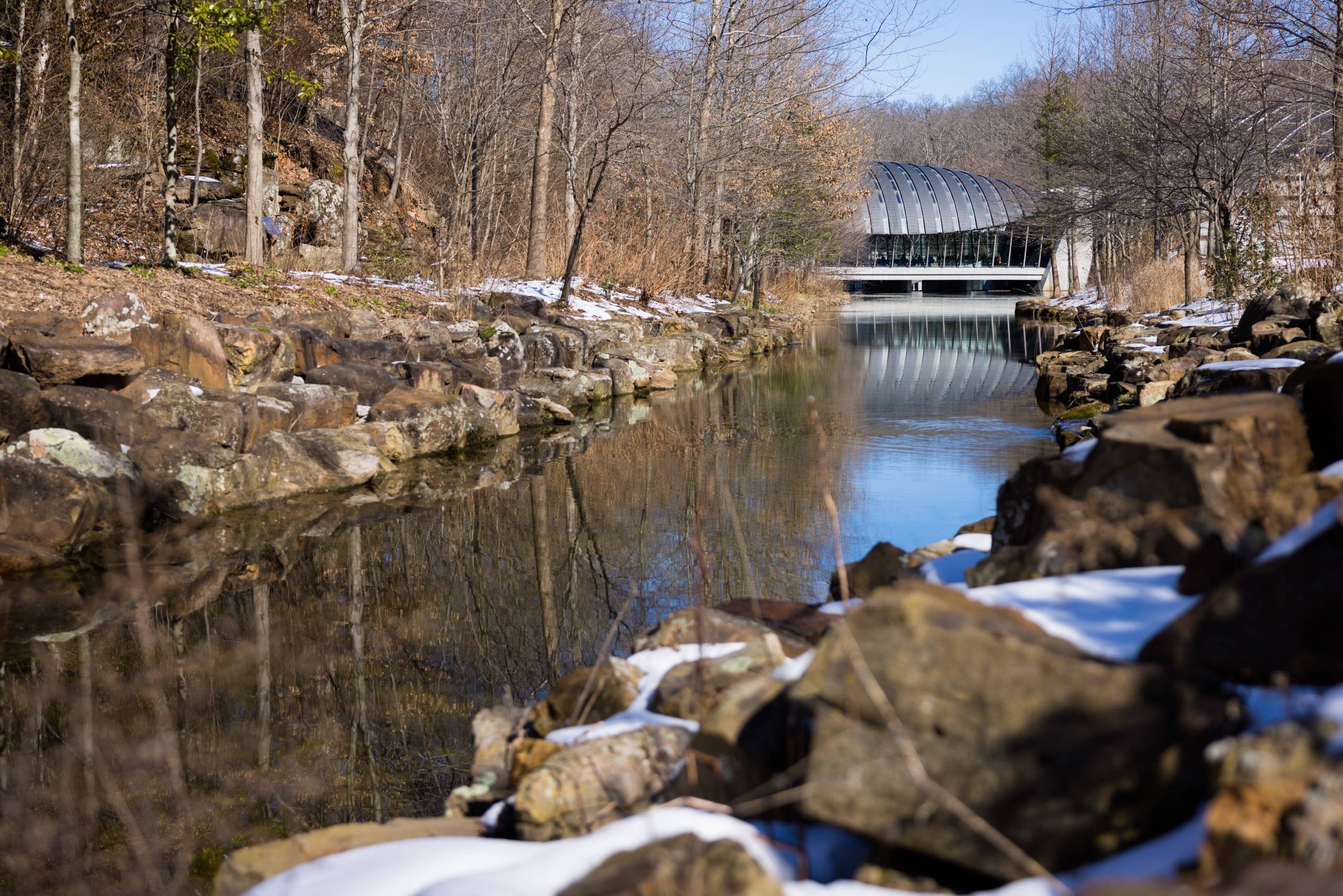 an exterior view of crystal bridges' eleven restaurant building from upstream surrounded by a winter landscape of trees, rocks, and snow on both sides