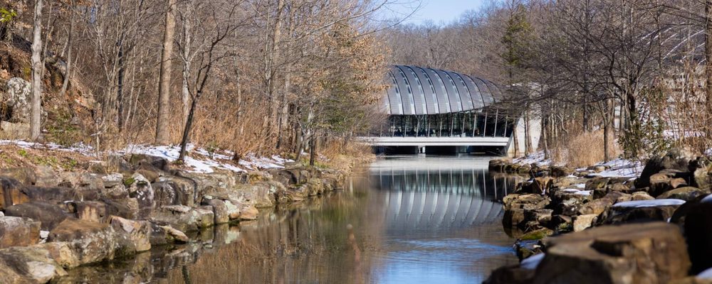 an exterior view of crystal bridges' eleven restaurant building from upstream surrounded by a winter landscape of trees, rocks, and snow on both sides