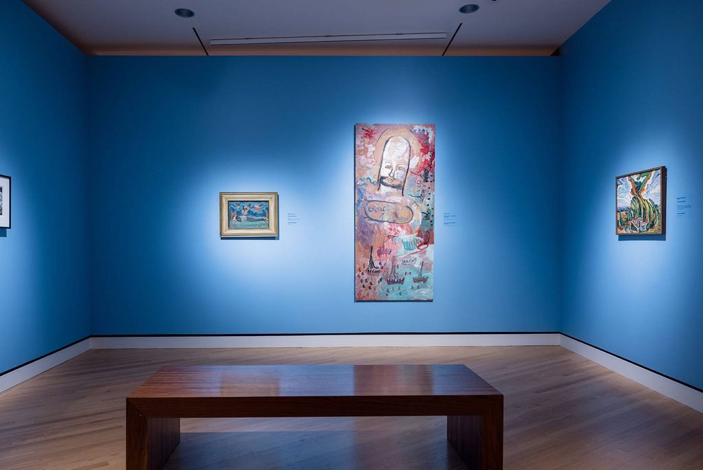 a gallery space featuring three painted works by Black artists against a bright blue wall and foregrounded by a viewing bench