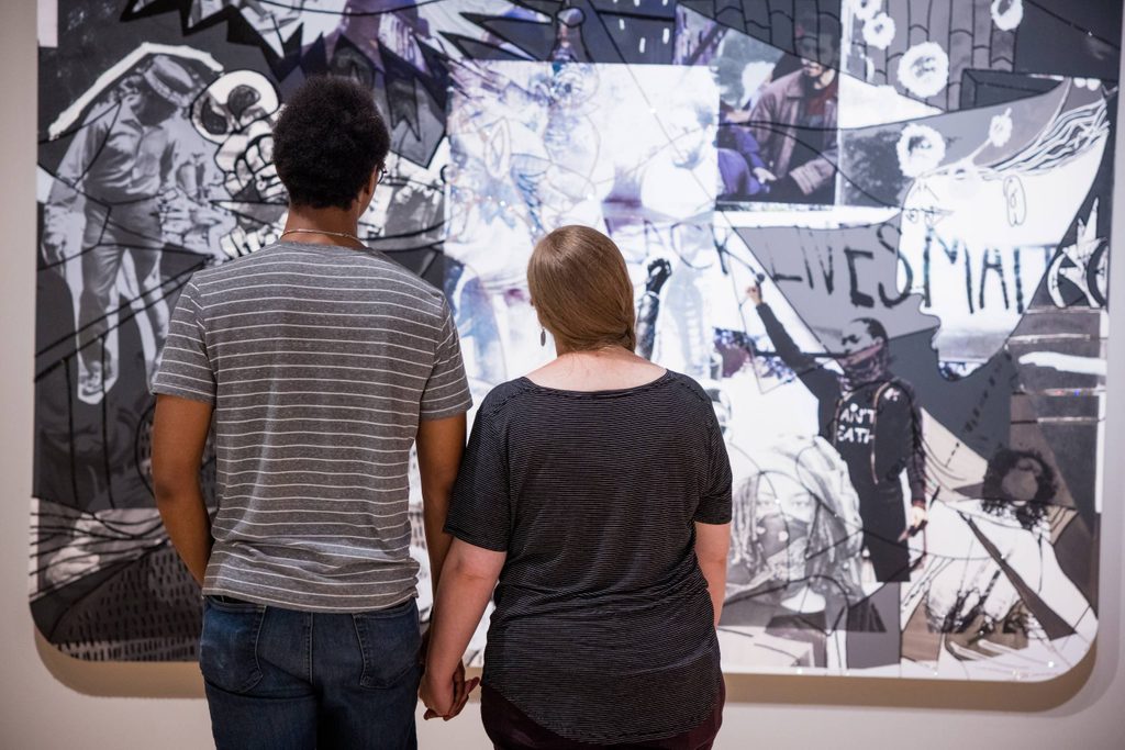 two people holding hands look at a black and white collage work featuring images of activists and phrases such as 