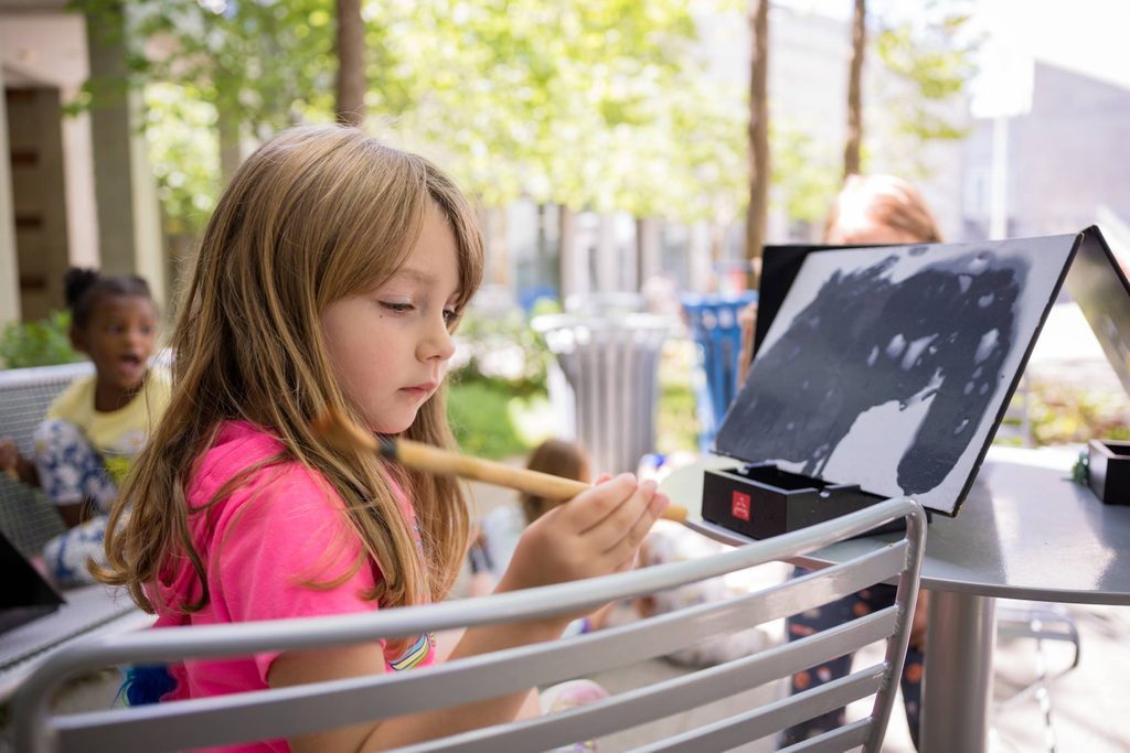 Young girl painting at a metal table outdoors in a pink shirt, with another child in the background.