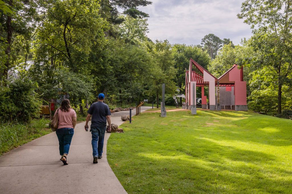 Two people walk along a paved trail lined with trees and grass. A small and angular pink house and other structures are visible among the trees ahead of them.