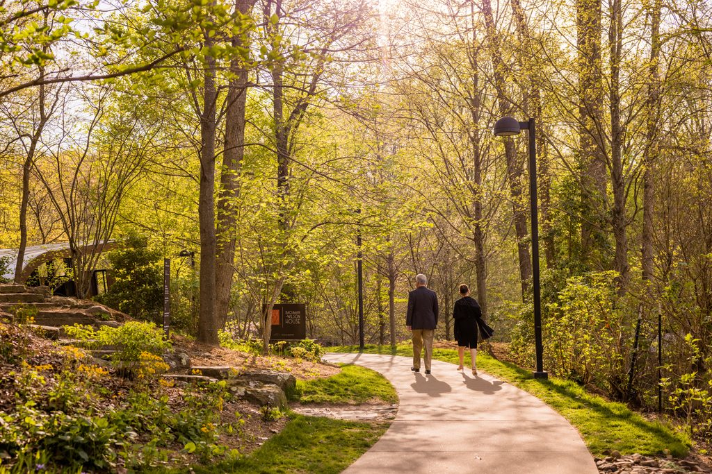 Couple walks on forest path near Bachman-Wilson House sign, sunlight filtering through trees.