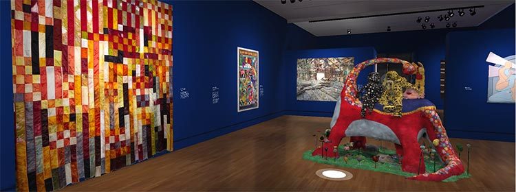 Art gallery with blue walls, large colorful quilt, framed artworks, and vibrant floor sculpture.