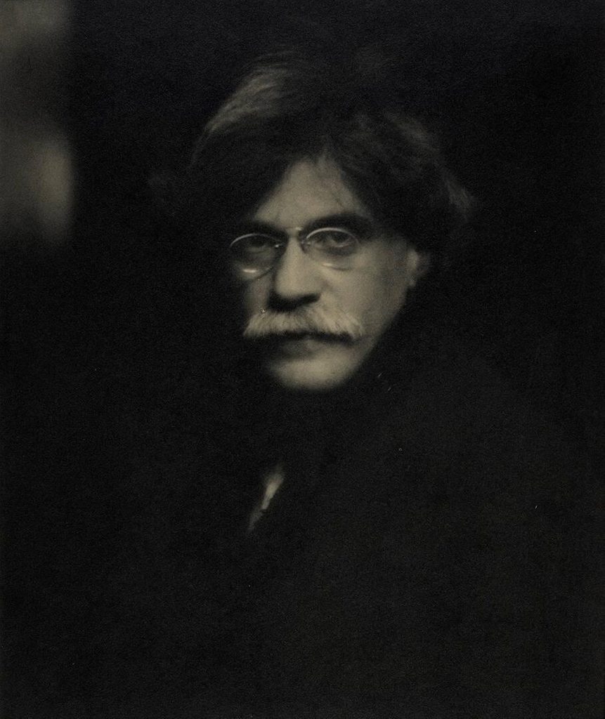Black and white photo of Alfred Stieglitz with thick white mustache, dark hair, wearing glass in front of dark background
