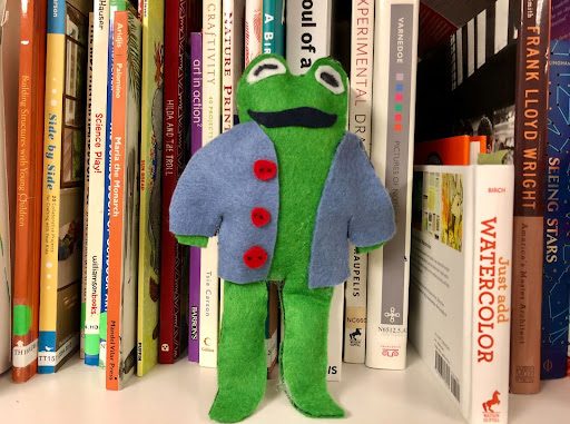 a green frog made out of felt wearing a blue jacket with red buttons in front of a shelf of books