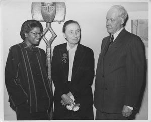 Three people stand together; one holds flowers; abstract art in background; black-and-white photo.