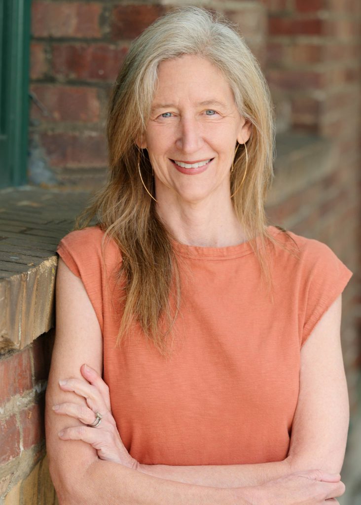 Middle-aged woman with long graying hair wearing orange top and hoop earrings leaning on brick wall.