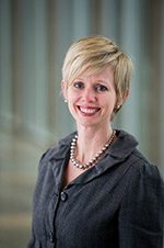 Person with short blonde hair in dark jacket and pearl necklace smiling at camera