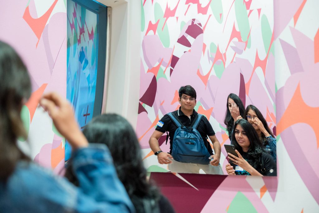 four young people look at their reflection in a funhouse mirror and hold up a camera