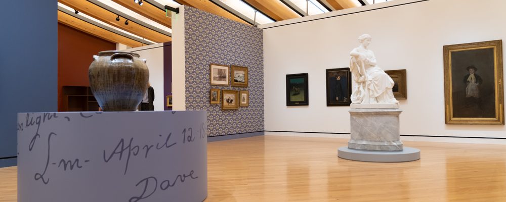 Art gallery interior with wooden floor, beams, paintings, a large urn, and a white marble statue.