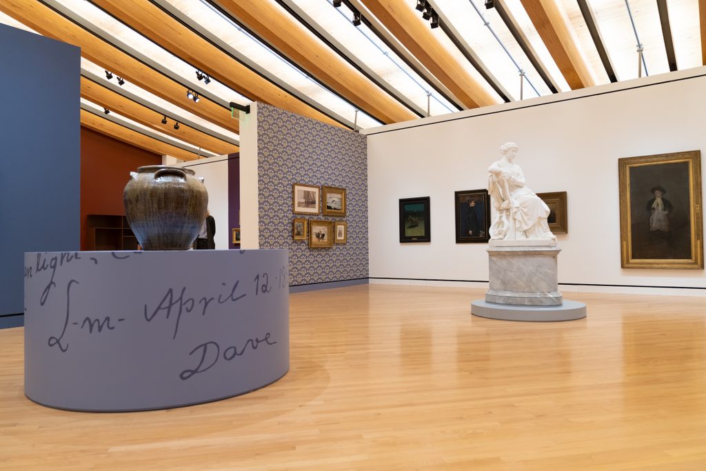 Art gallery interior with wooden floor, beams, paintings, a large urn, and a white marble statue.