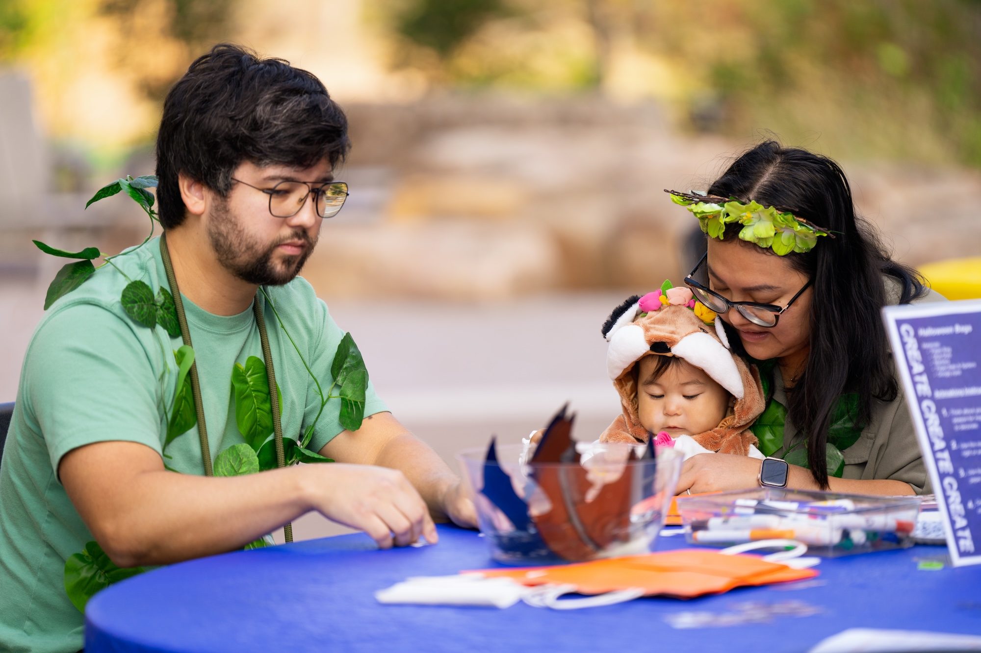 Adults with foliage accessories and child in costume crafting at blue table with art supplies.