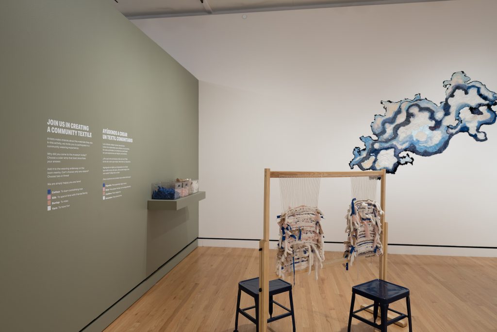 Interactive community textile exhibit with loom, stools, materials, and abstract artwork on wall.