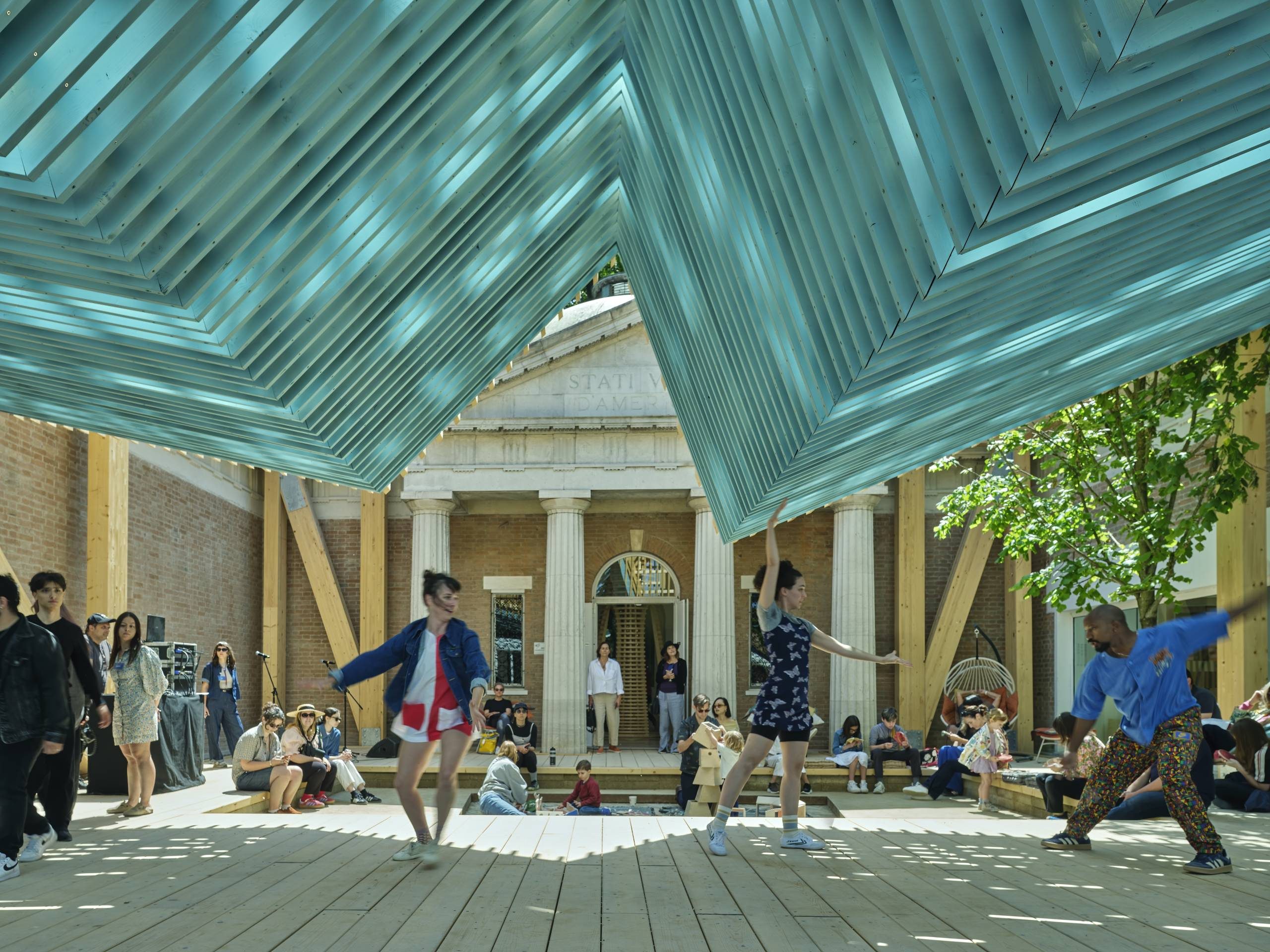 Outdoor wooden stage with dancers beneath geometric overhead structure and seated spectators