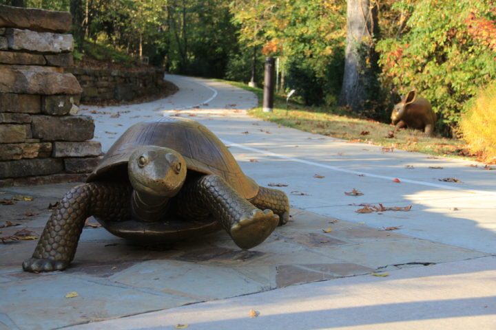 Bronze turtle statue on a paved path with a bronze hare in the background in a lush, green landscape.