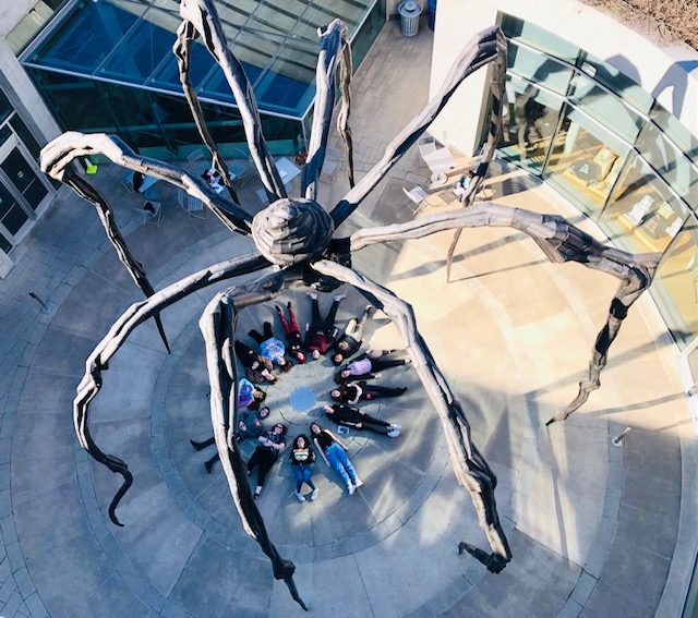 a group of student visitors lay in a circle underneath maman in the museum courtyard