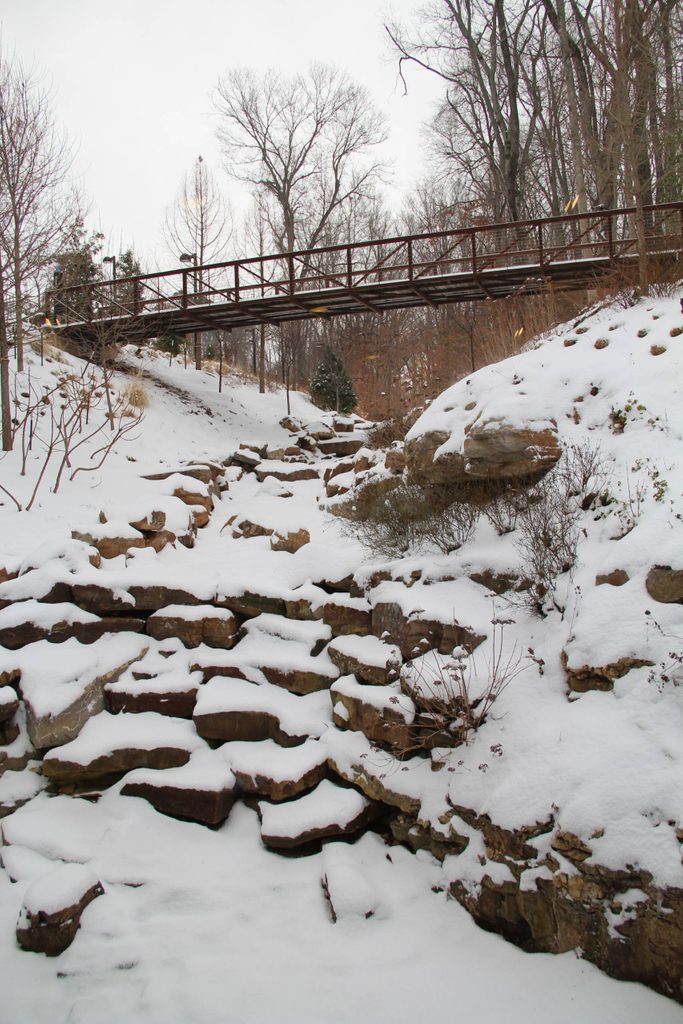 Snowy landscape with stone path and rusty bridge surrounded by leafless trees.