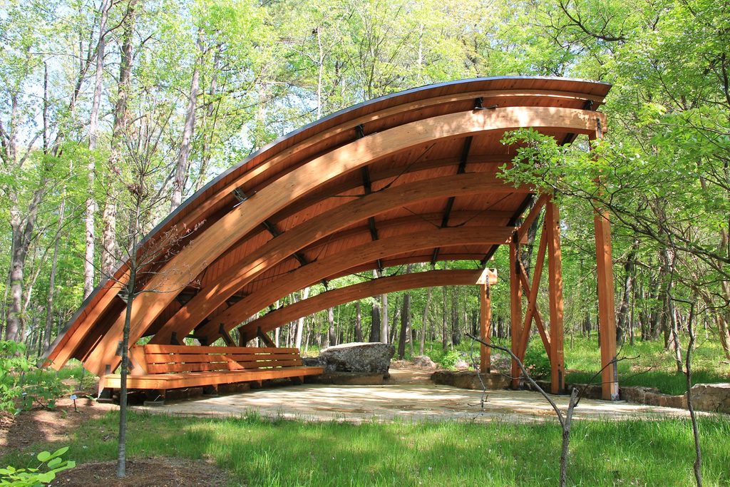 Curved wooden pavilion in a wooded area with benches and greenery.