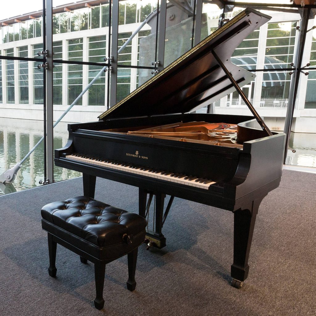 Grand piano with open lid before large window overlooking water; black leather bench in front.