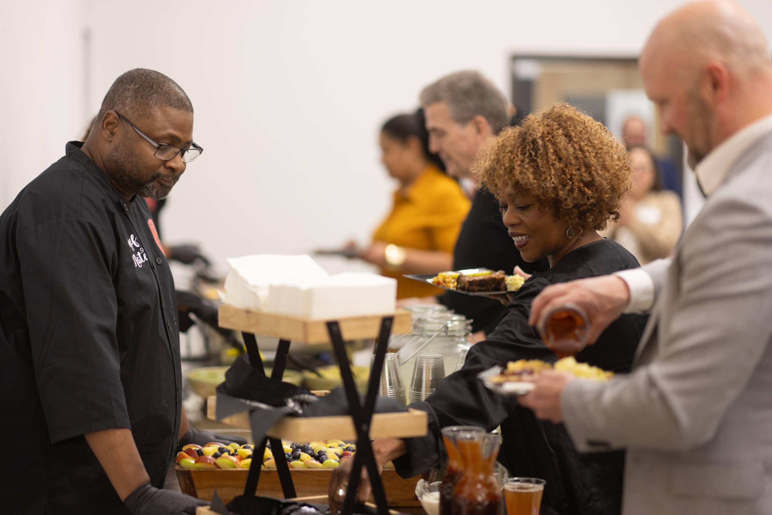 People enjoy buffet, served by a male server in black, with drinks and assorted dishes nearby.