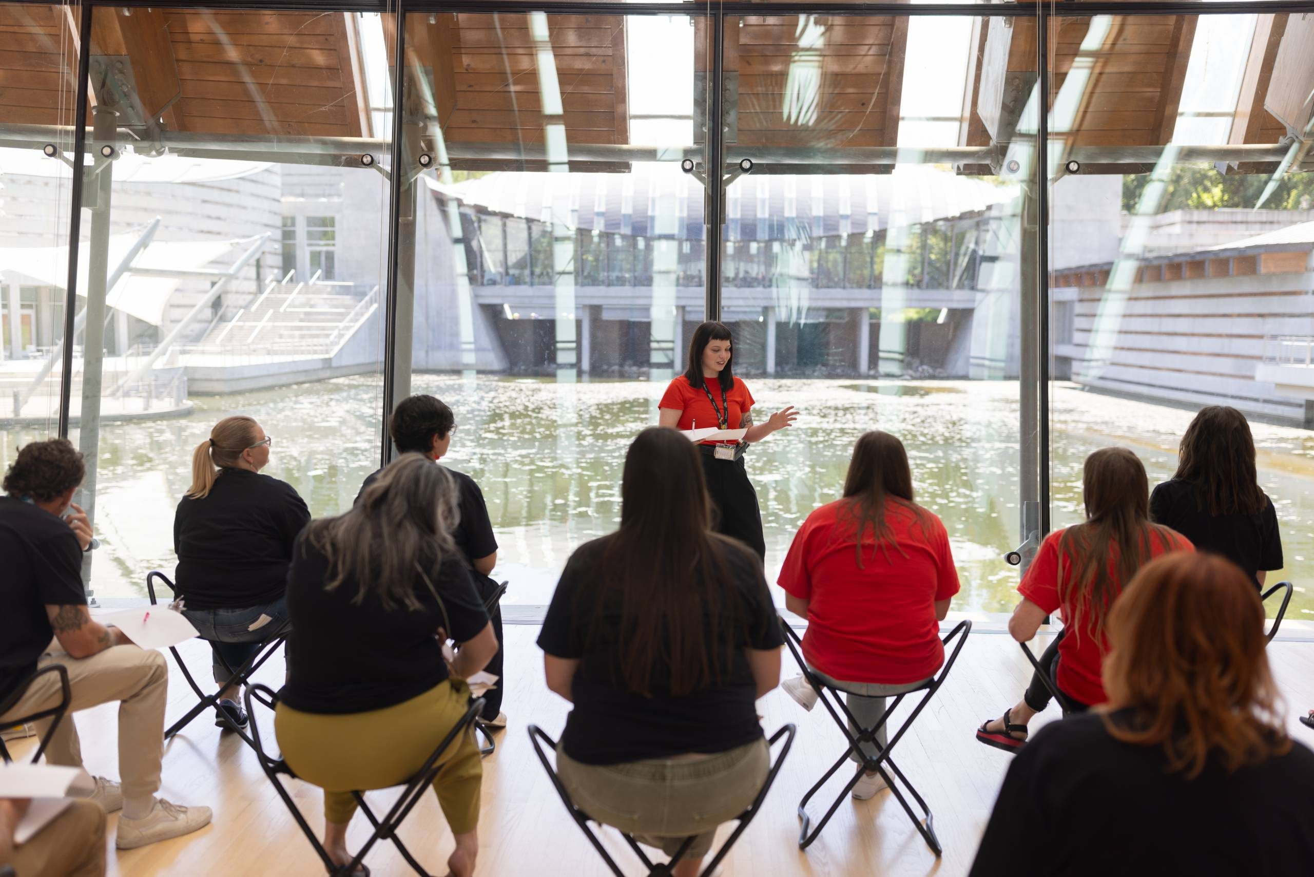 Group listening to a speaker in a glass-walled room overlooking a pond and building outside.