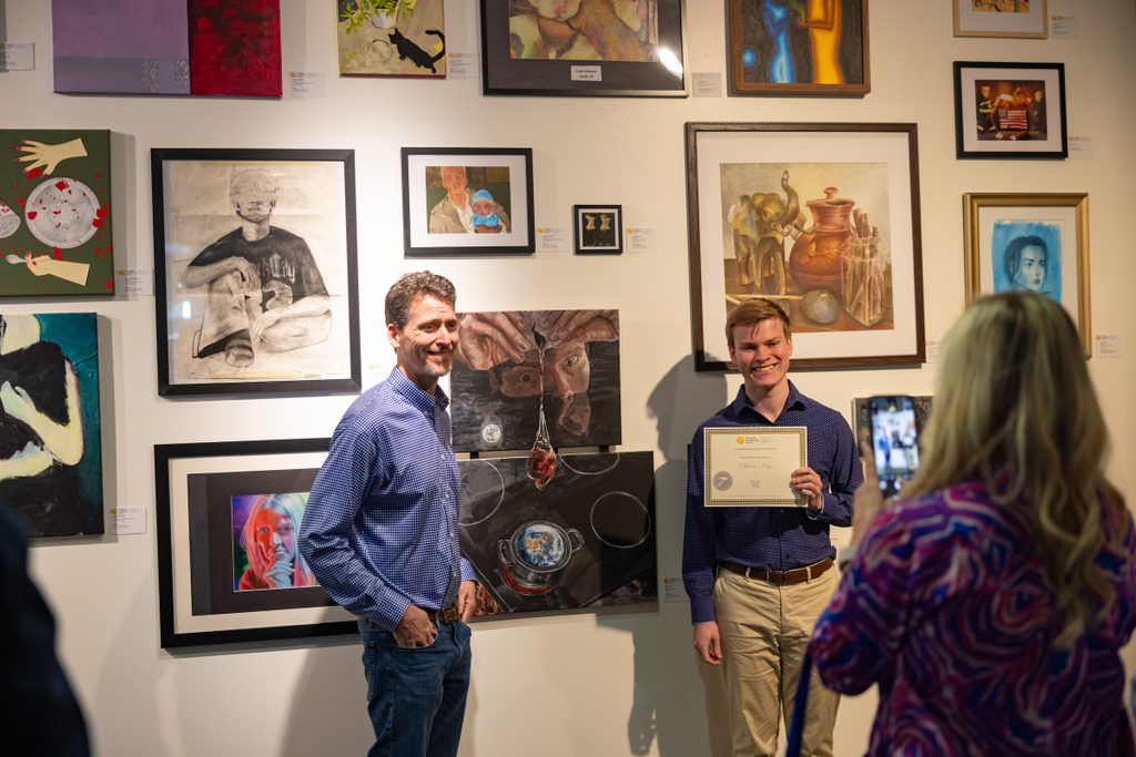 A young man holding a certificate stands next to an older man in front of framed artworks.